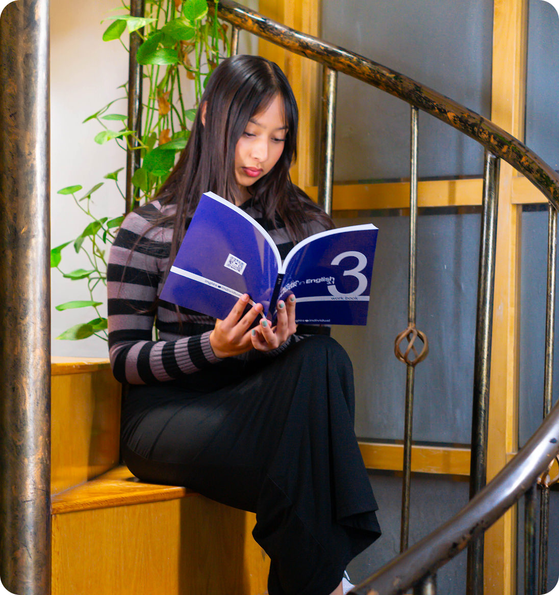Woman sitting on wooden stairs reading a blue workbook near a metal spiral railing and green plants.