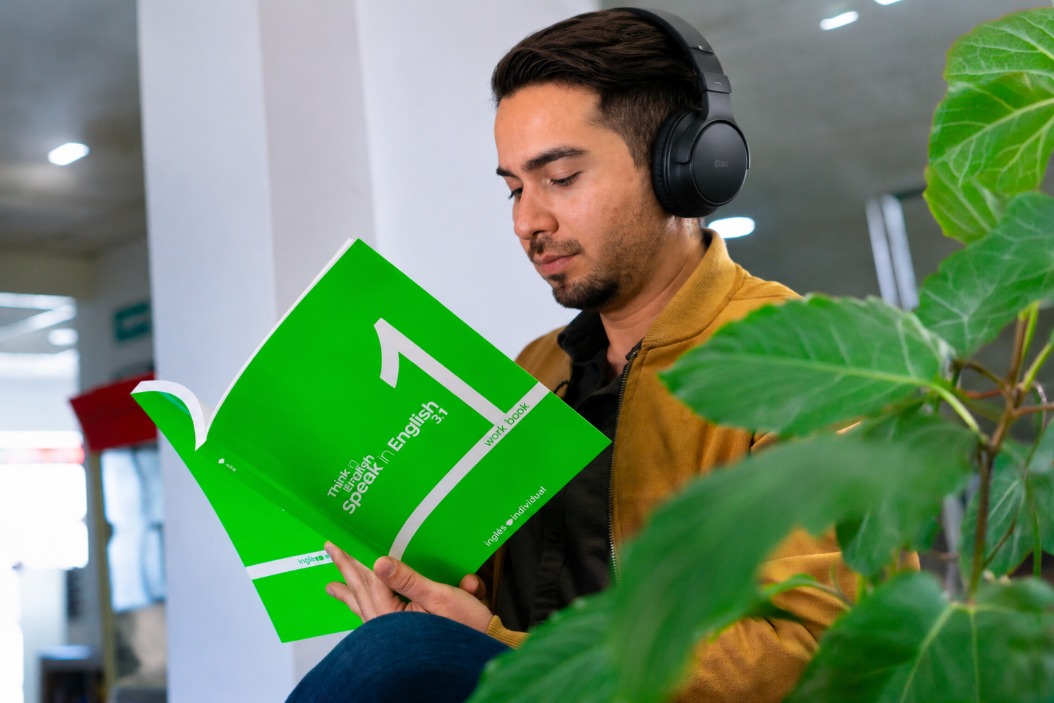 Man wearing headphones reading a green workbook titled 'Speak in English 3.1' indoors.