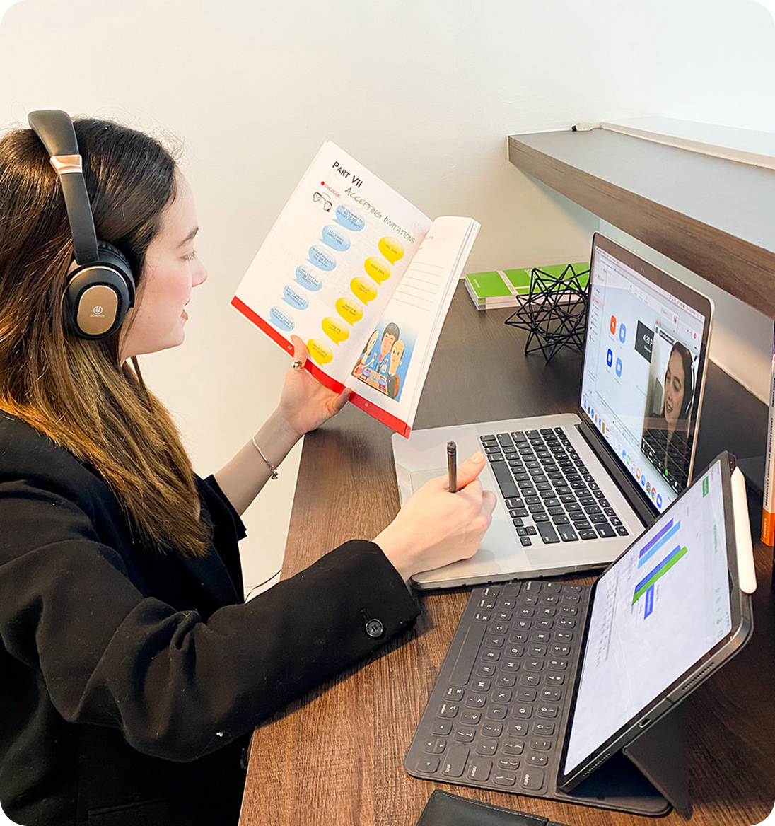 Young woman wearing headphones studies with book, laptop showing video call, and tablet with data chart.